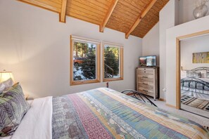 Une chambre à coucher comprenant un lit, une commode, un téléviseur et deux fenêtres donnant sur des arbres et une voiture.