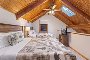 A bedroom with a sloped ceiling, a bed with a quilt, a nightstand, a lamp, a TV on a wooden stand, and a window with a view. - Riverside Sanctuary • RMNP • River Views (Estes Park)
