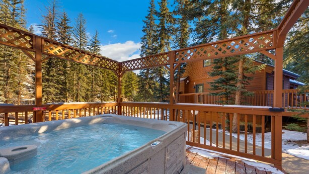 A hot tub in a wooden deck area with a wooden pergola overhead, surrounded by trees and a cabin in the background. - Riverside Sanctuary • RMNP • River Views (Estes Park)