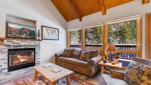 A cozy living room with a fireplace, a sofa, a coffee table, and large windows offering a view of the outdoors. - Riverside Sanctuary • RMNP • River Views (Estes Park)