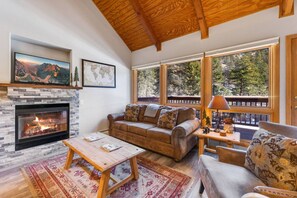 A cozy living room with a fireplace, a sofa, a coffee table, and large windows offering a view of trees. - Riverside Sanctuary • RMNP • River Views (Estes Park)