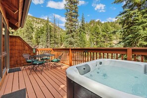 A wooden deck with a hot tub, outdoor seating, and a view of a forested mountain. - Whispering River Escape • Private Hot Tub • RMNP (Estes Park)