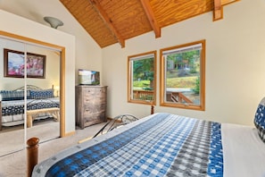 A bedroom with a bed, a dresser, a television, and a window with a view of a garden. - Whispering River Escape • Private Hot Tub • RMNP (Estes Park)