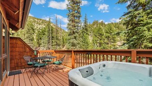 A wooden deck with a hot tub, outdoor seating, and a view of a forested mountain. - Whispering River Escape • Private Hot Tub • RMNP (Estes Park)