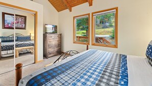 A bedroom with a large bed, a dresser, a television, and a view of the outdoors through two windows. - Whispering River Escape • Private Hot Tub • RMNP (Estes Park)