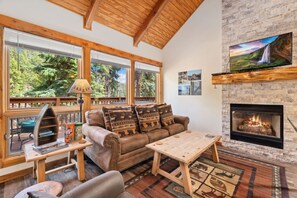 A cozy living room with a fireplace, wooden furniture, and large windows overlooking a forest. - Whispering River Escape • Private Hot Tub • RMNP (Estes Park)