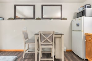 Eine kleine Küchenzeile mit einem weißen Kühlschrank, einer Mikrowelle und einer kleinen Arbeitsfläche. Zwei Stühle und ein Tisch bieten Platz, und der Boden ist aus Holz.