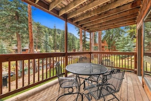 A covered outdoor deck with a table and chairs, surrounded by a wooden railing, overlooking a forested area.