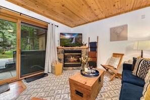Una sala de estar acogedora con chimenea, televisor y un amplio ventanal con vistas a un bosque.