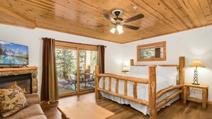A cozy room with a wooden ceiling, a fireplace, a bed with a wooden frame, a table, and a view of the outdoors. - Updated Riverfront Retreat • A/C • Private Hot Tub (Estes Park)
