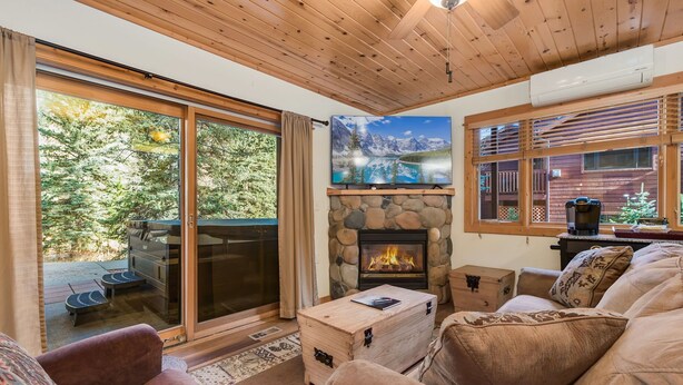 A cozy living room with a stone fireplace, a large TV, and a view of a forested area. - Updated Riverfront Retreat • A/C • Private Hot Tub (Estes Park)