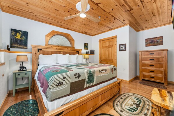 A bedroom with a wooden ceiling, a bed with a quilt, a nightstand, a dresser, and a door. - River Retreat • River Views • Near RMNP (Estes Park)