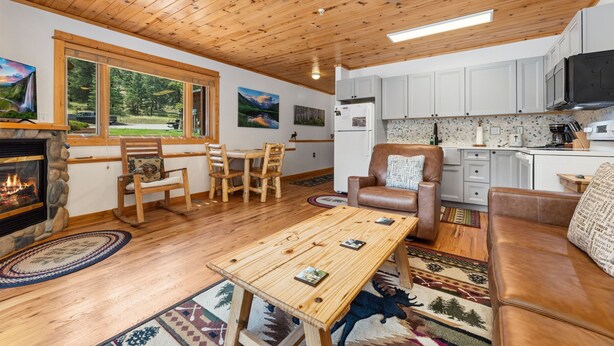 A cozy living room with a fireplace, wooden furniture, a dining area, and a kitchen with white cabinets. - River Retreat • River Views • Near RMNP (Estes Park)