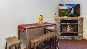 A cozy room with a stone fireplace, a wooden table with a red and black checkered tablecloth, and a television mounted above the fireplace displaying a scenic landscape.