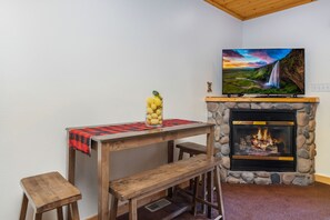 A cozy room with a stone fireplace, a wooden table with a red and black checkered tablecloth, and a television mounted above the fireplace displaying a scenic landscape.