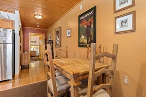 A wooden dining table with chairs, a poster on the wall, and a kitchen area with a refrigerator.