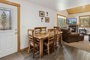 A dining area with a wooden table and chairs, a fireplace, and a sofa in a room with wooden beams and large windows. - Fall River Retreat • Private Hot Tub (Estes Park)