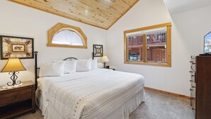 Quarto com teto de madeira, cama grande com roupa de cama branca, duas luminárias de cabeceira, cômoda de madeira e janela com vista para um prédio de tijolos.