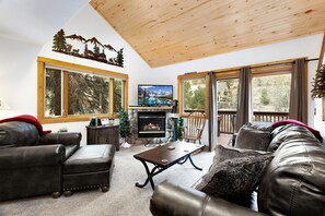 A cozy living room with a fireplace, leather sofas, a wooden ceiling, and large windows with a view of trees. - Quiet River Escape • Private Hot Tub • Close to RMNP (Estes Park)