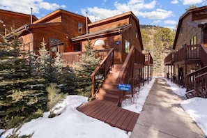 A wooden cabin with a staircase leading to a deck, surrounded by snow and evergreen trees.