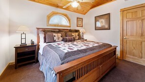 Un dormitorio con techo de madera, una cama con colcha estampada, dos mesitas de noche con lámparas, una ventana con diseño de sol y una puerta de madera.
