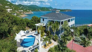 Une maison à deux étages avec une grande piscine, entourée d’une végétation luxuriante et offrant une vue sur l’océan.