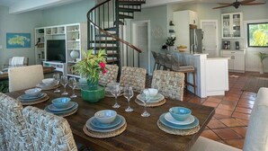 Un coin repas avec une table en bois dressée pour six personnes, entourée de chaises en osier. Un escalier en colimaçon est visible au fond, menant à un niveau supérieur.
