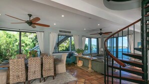 Un vaste espace de vie comprenant un coin repas, un escalier et de grandes fenêtres donnant sur un paysage verdoyant, ainsi qu’un balcon.