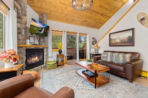 A cozy living room with a stone fireplace, a leather sofa, a coffee table, and a large window offering a view of the outdoors. - Cozy Riverfront Hideaway • Private Hot Tub (Estes Park)