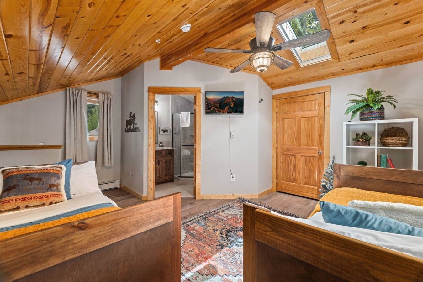 A room with a wooden ceiling, a bed with a patterned pillow, a small table, a wall-mounted TV, and a door leading to a bathroom.