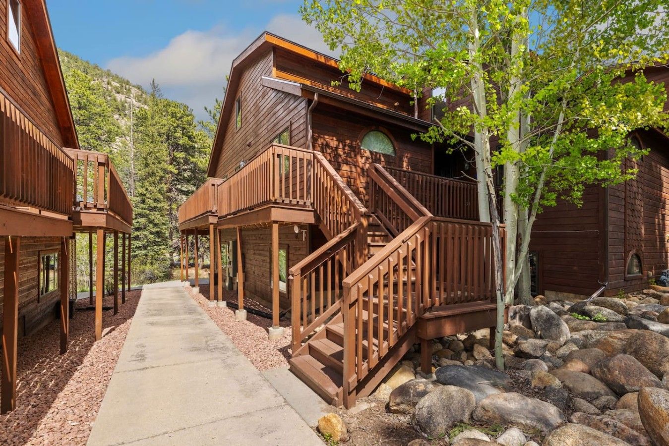 A wooden cabin with a staircase leading to a deck, surrounded by trees and rocks.