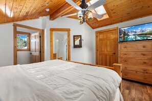 Un dormitorio con una cama grande, techo de madera, ventilador de techo, cómoda y un televisor que muestra un paisaje montañoso.