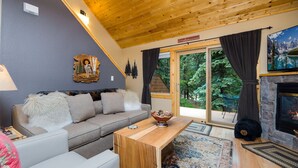 Un salon chaleureux avec un plafond en bois, une cheminée en pierre, un canapé, une table basse et une vue sur une forêt, visible à travers des portes vitrées.