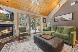 A cozy living room with a fireplace, a green sofa, a leather ottoman, and a large window offering a view of the outdoors. - Fall River Hideaway • Private Hot Tub • Near RMNP (Estes Park)