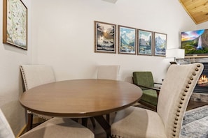 A dining area with a round table and four chairs, a fireplace with a painting above it, and a wall with three framed posters. - Fall River Hideaway • Private Hot Tub • Near RMNP (Estes Park)