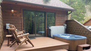 A wooden cabin with a hot tub, a deck with chairs, and a sliding glass door leading to a covered outdoor area. - Fall River Hideaway • Private Hot Tub • Close to RMNP (Estes Park)