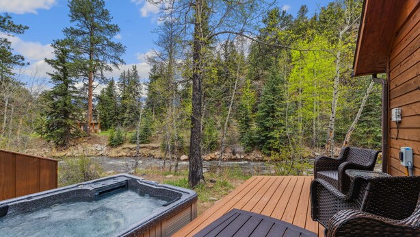 Une terrasse en bois avec un jacuzzi et des meubles en osier, entourée d’une forêt et d’une rivière.