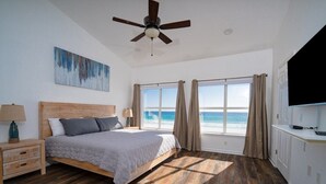 Un dormitorio con piso de madera, una cama con ropa de cama gris, un ventilador de techo, un cuadro en la pared y vista al océano a través de grandes ventanales.