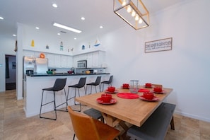 Una cocina moderna con armarios blancos, una isla central con taburetes altos y una mesa de comedor para cuatro personas, con vajilla roja.