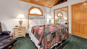 Une chambre avec un plafond en bois, un lit recouvert d’une courtepointe, deux tables de chevet avec des lampes, une chaise et une porte en bois.
