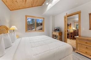 A bedroom with a bed, a wooden dresser, a desk with a chair, a window with a view of trees, and a mirror.