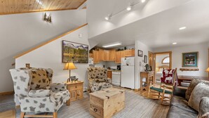 A cozy living room with a sofa, armchair, and a wooden chest. A kitchen is visible in the background with wooden cabinets and a refrigerator. - Fall River Retreat • Mountain Views • RMNP (Estes Park)