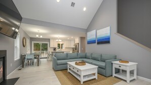 En moderne stue med en hjørnesofa, et sofabord og et lille bord. Der er en pejs til venstre, et køkken i baggrunden og et spiseområde med stole og bord.