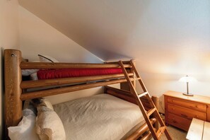 A bunk bed with a wooden ladder, a bedside table with a lamp, and a small wooden dresser.