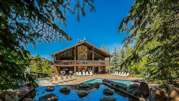 Una cabaña con piscina rodeada de rocas, una terraza con sillones de descanso y un fondo de bosque.
