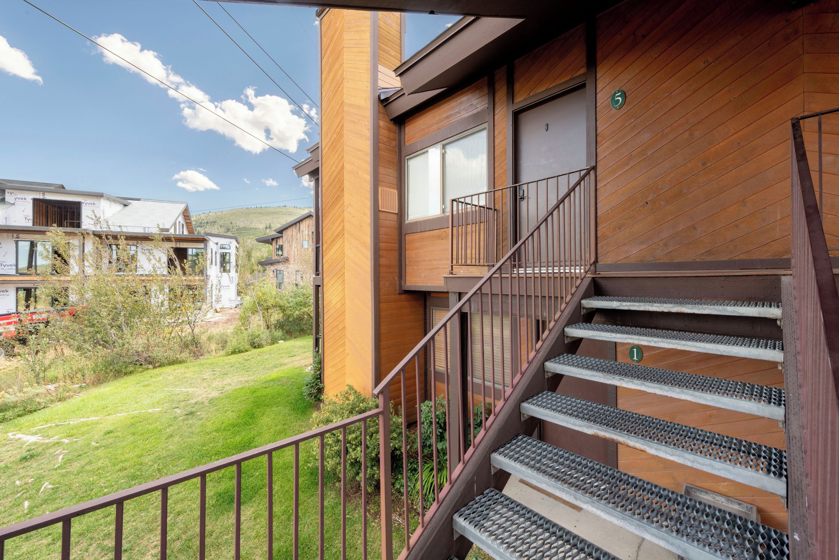A residential building with a wooden exterior, a staircase with metal railings, and a view of a grassy area with other buildings in the background.