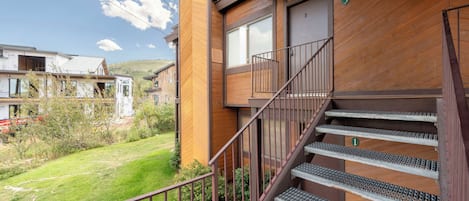 A residential building with a wooden exterior, a staircase with metal railings, and a view of a grassy area with other buildings in the background.