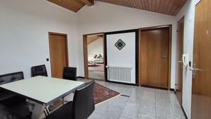 Une pièce avec une table blanche, un canapé en cuir noir et un tapis rouge à motifs. On y trouve deux portes en bois et un radiateur blanc.