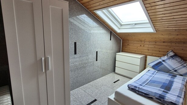 Une chambre avec un puits de lumière, une armoire, un lit avec une couette à carreaux et une petite table de chevet.