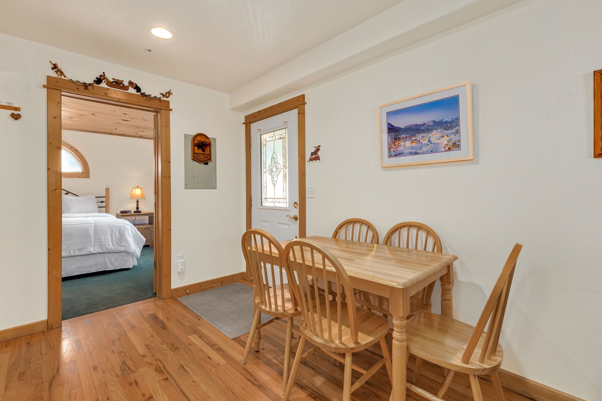 A dining area with a wooden table and chairs, a bed in the adjacent room, and a framed picture on the wall.